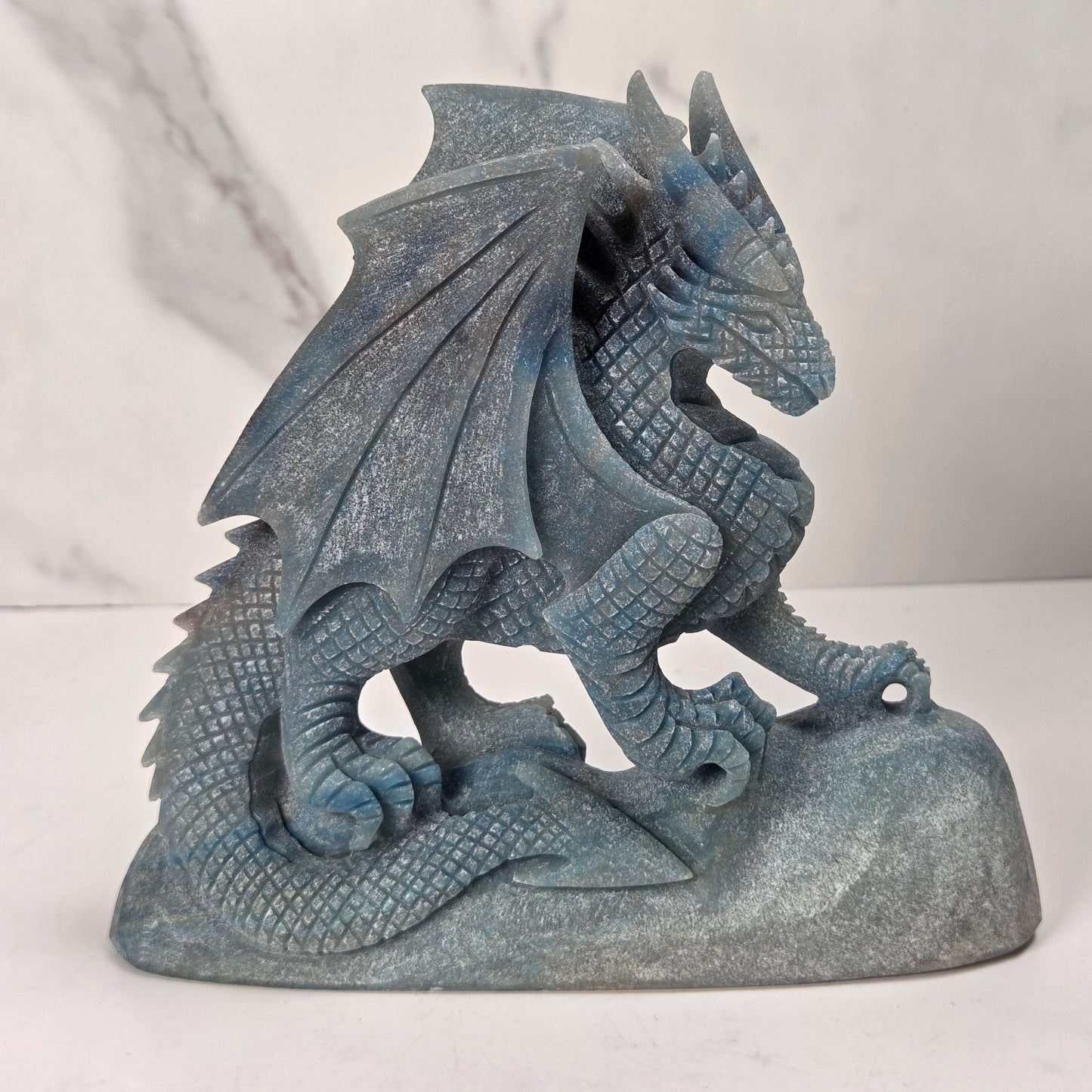 A blue-gray stone dragon figurine standing on a rock base, photographed against a light marble-patterned background.