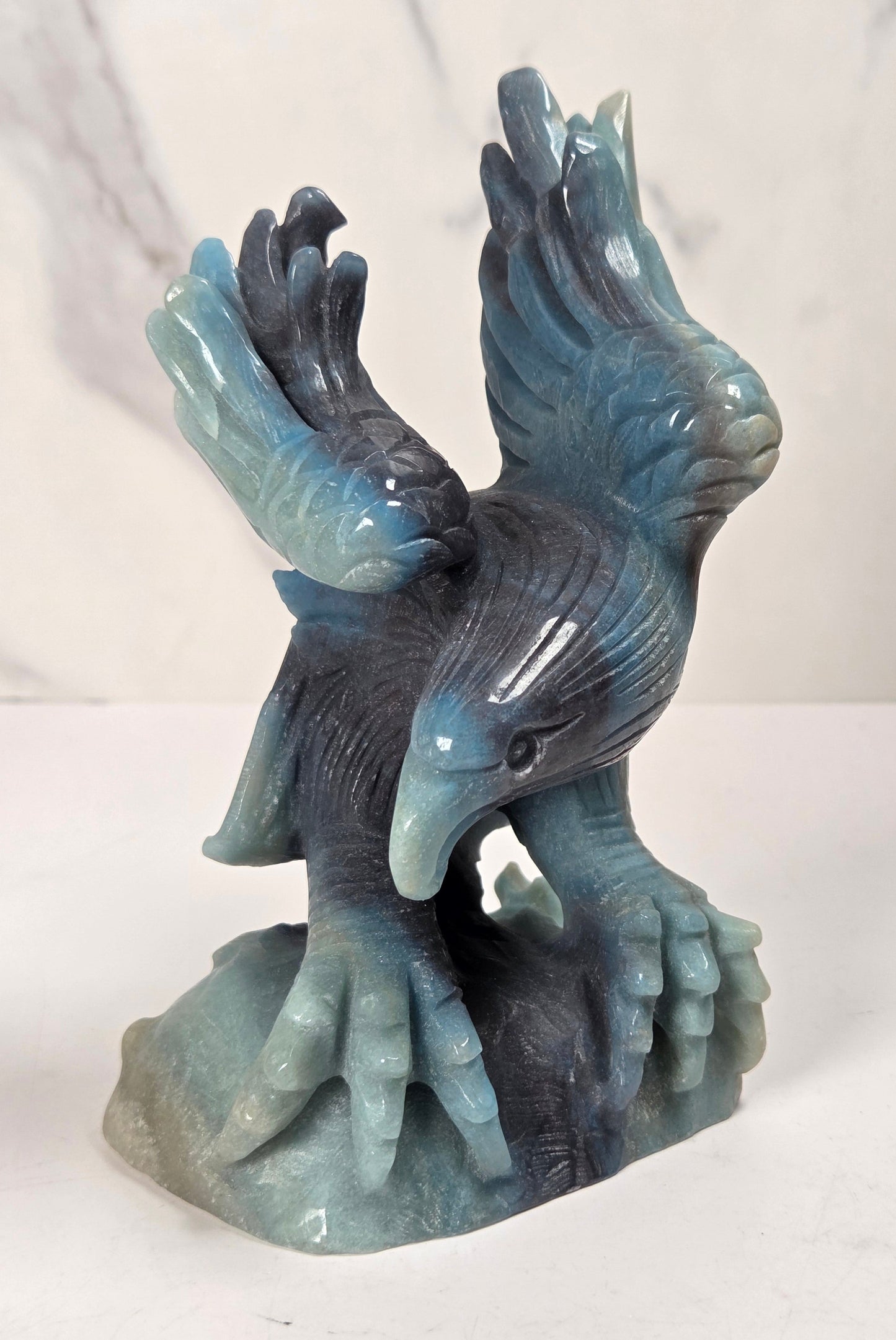 A carved stone sculpture of a bird of prey with wings spread, standing on a rocky base. The material has a marbled blue, gray, and teal coloration, resembling polished slate or a similar stone.