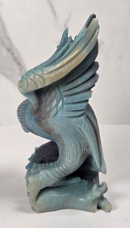 A small sculpted winged creature figurine in a pale blue-gray stone-like finish, standing on a white surface against a light gray marble-pattern backdrop.