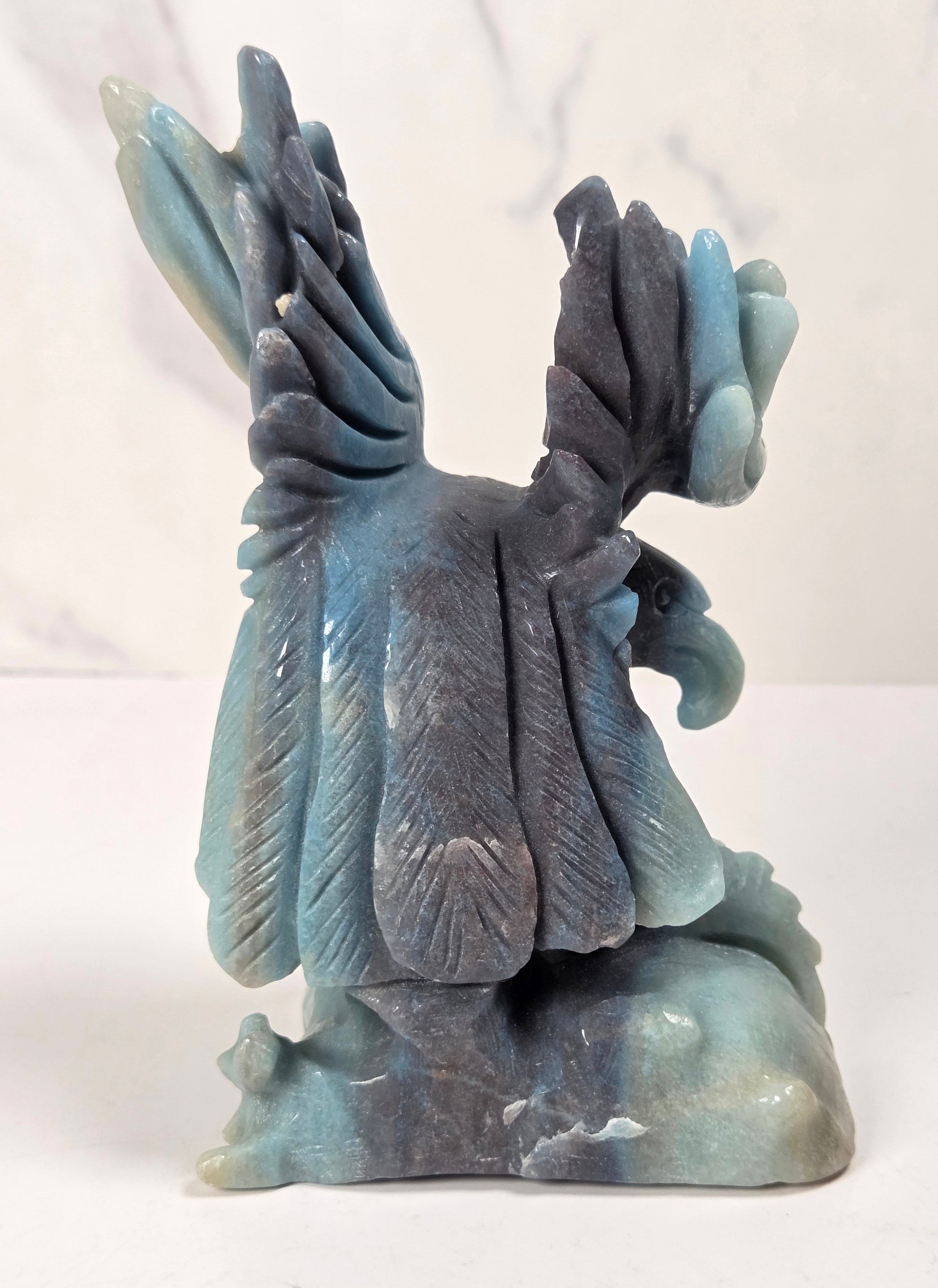 A carved stone sculpture of a bird with wings spread, featuring shades of blue, gray, and teal, set against a light marble-patterned background.