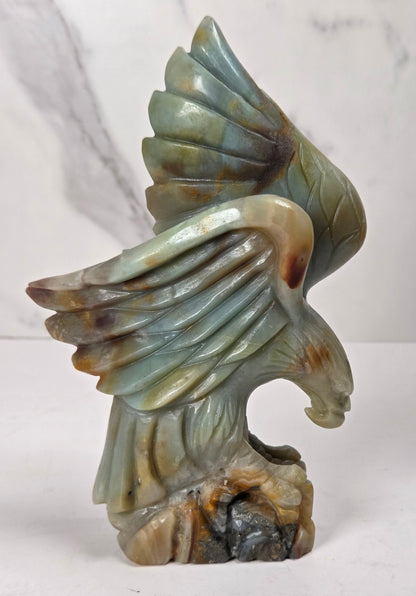 A carved stone eagle sculpture with wings spread, standing on a rocky base. The stone has a marbled coloration of pale green, beige, and brown, photographed against a light gray marble-patterned background.