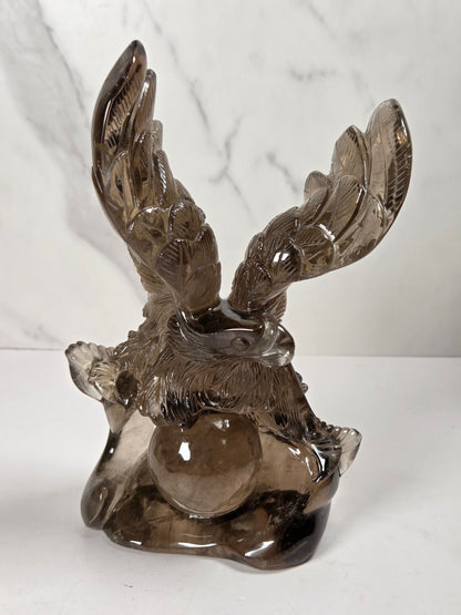 A smoky brown translucent glass sculpture of a winged figure, likely a cherub or angel, holding a glossy glass orb, set against a white marble-patterned background.