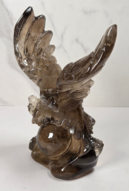 A smoky brown translucent glass eagle figurine with wings spread, holding a glowing glass orb, set against a white marble-patterned background.