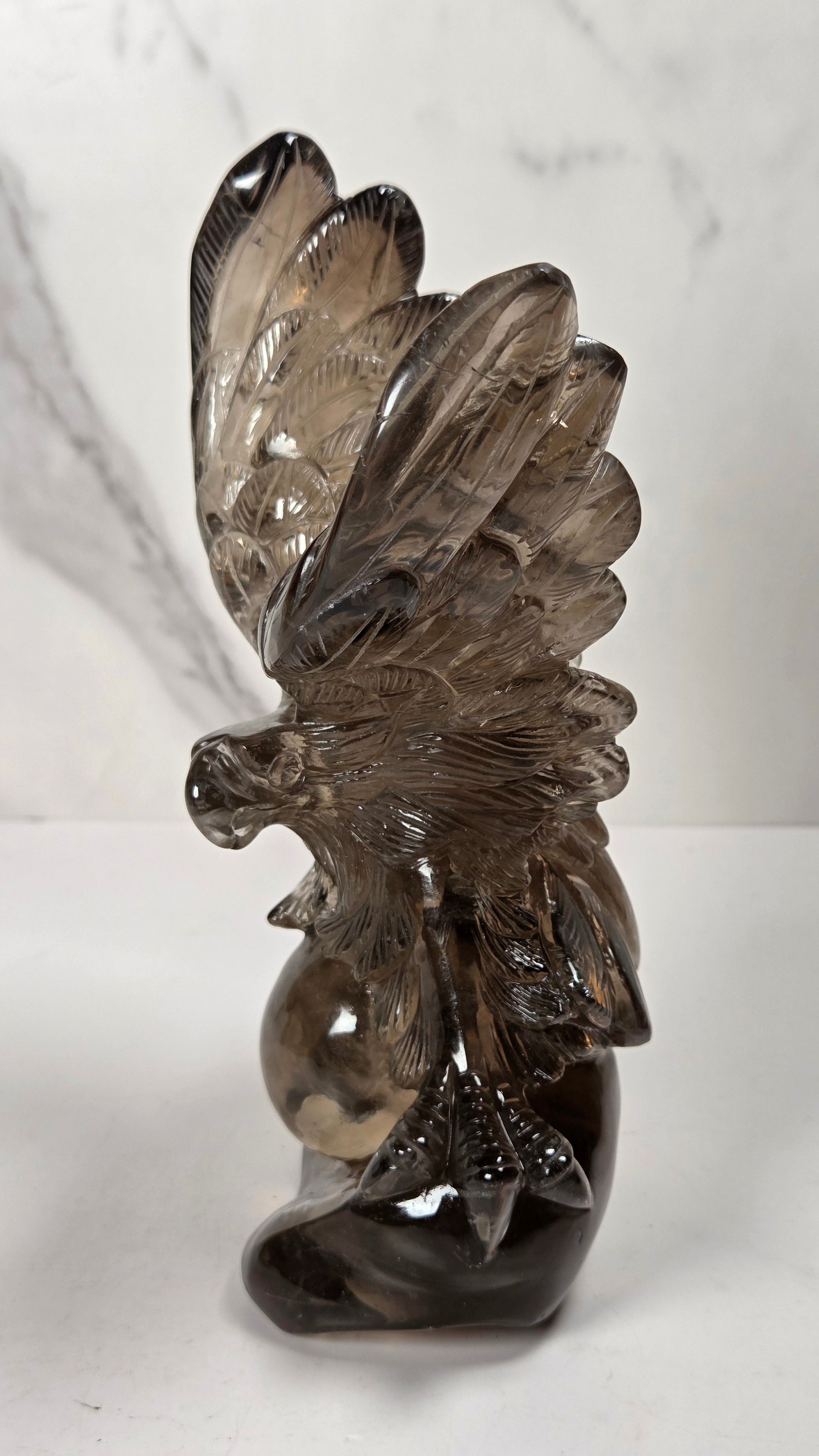 A smoky brown glass sculpture of a majestic eagle with wings spread wide, standing on a white surface against a white marble-patterned background.