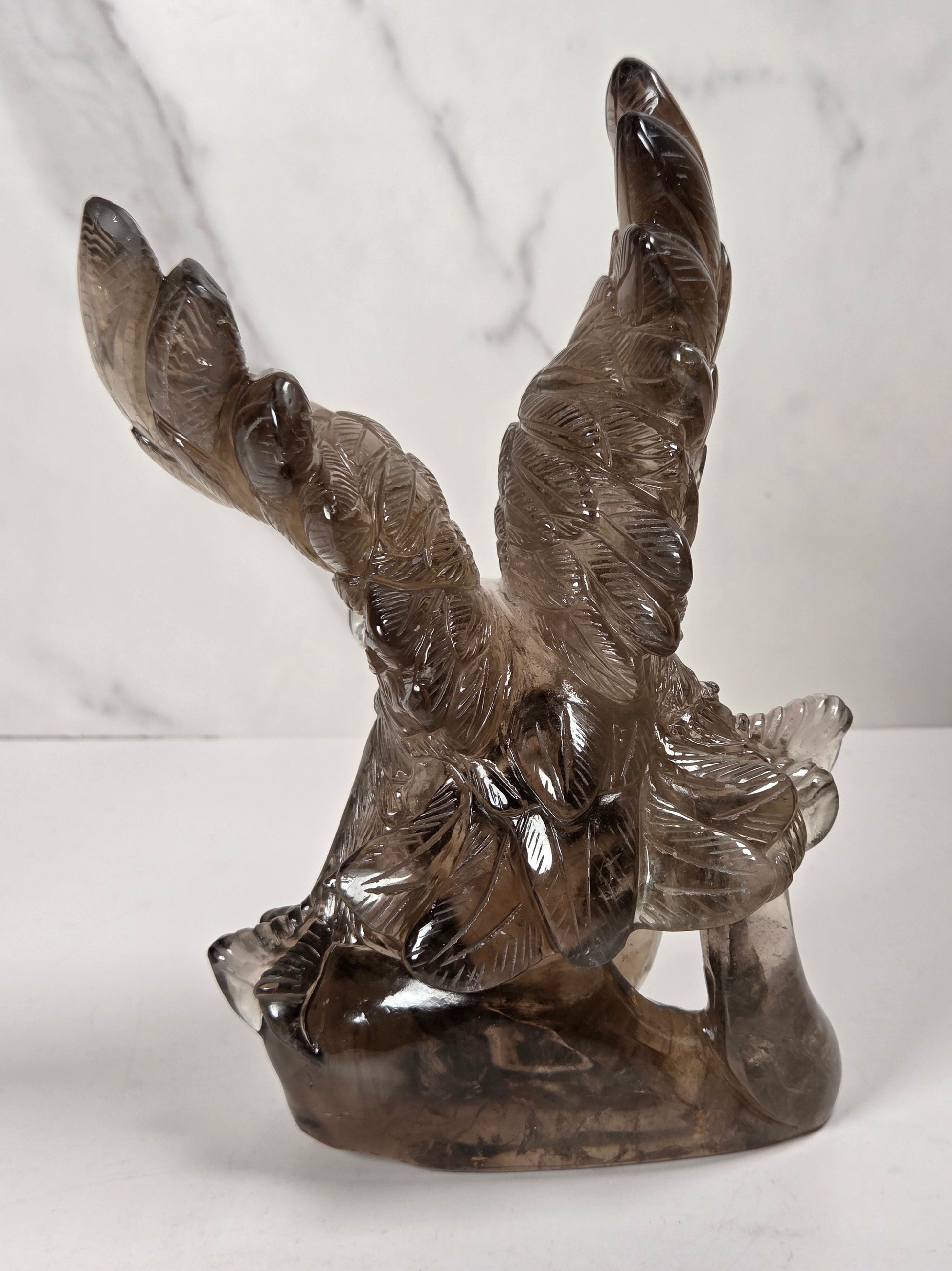 A smoky brown translucent glass sculpture of a winged figure, likely a cherub or angel, standing on a matching base against a white marble-patterned background.