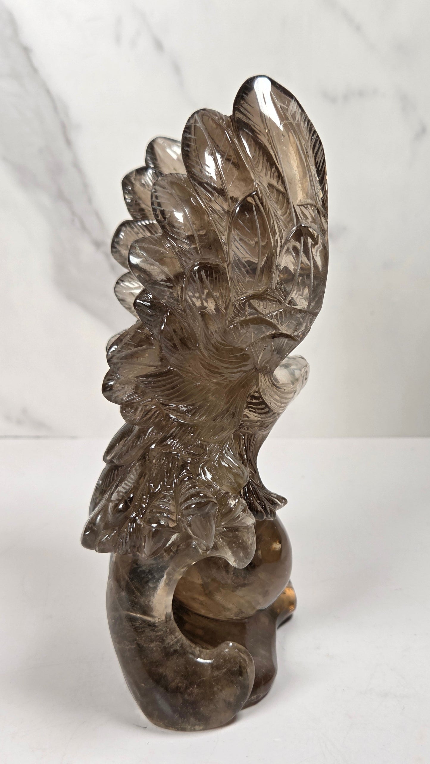 A carved smoky quartz bird sculpture, likely a phoenix or dragon, standing on a white surface against a light gray marble-patterned background.