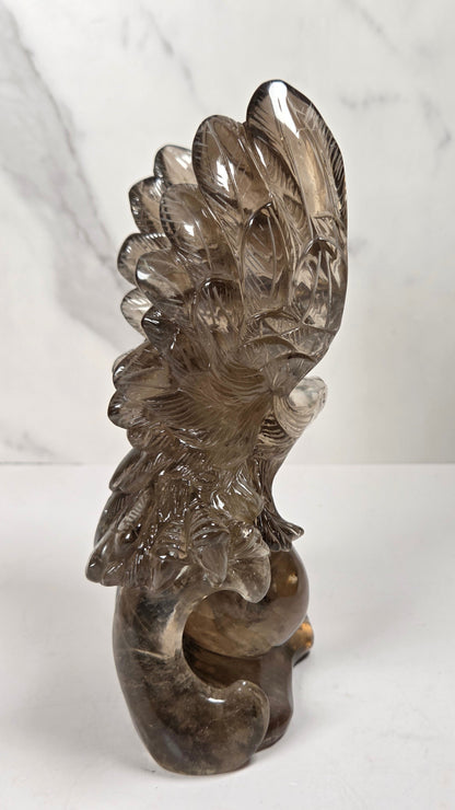 A carved smoky quartz bird sculpture, likely a phoenix or dragon, standing on a white surface against a light gray marble-patterned background.