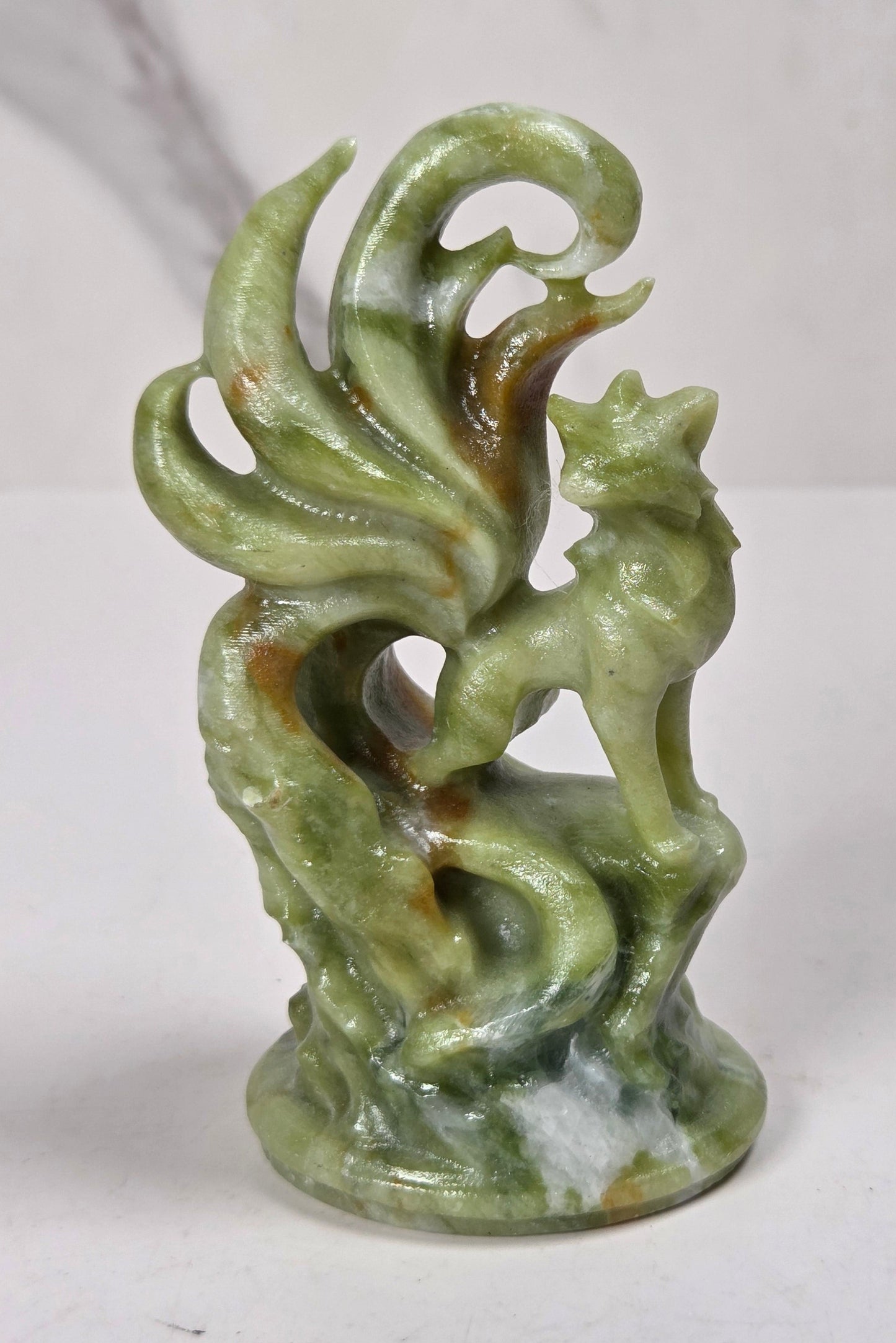 A small green jadeite-style figurine of a fox or fox-like creature with curled tail feathers or leaves, standing on a base, photographed against a light marble-patterned background.
