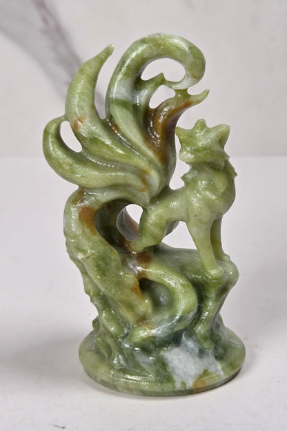 A small green jadeite-style figurine of a fox or fox-like creature with curled tail feathers or leaves, standing on a base, photographed against a light marble-patterned background.