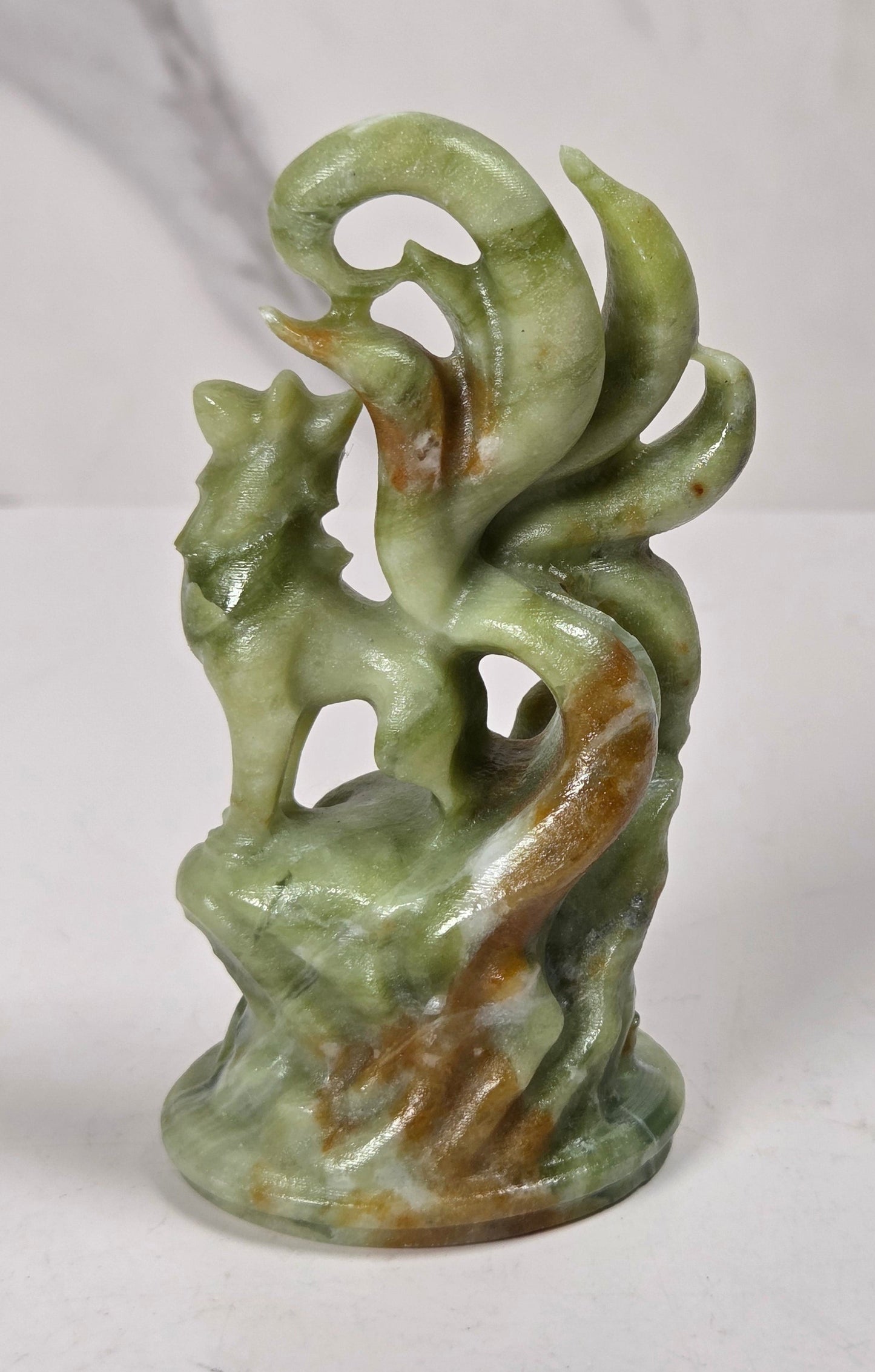 A small carved figurine of a fox or wolf standing on a rock-like base, made from polished green stone with brown marbling, photographed against a light marble-patterned background.