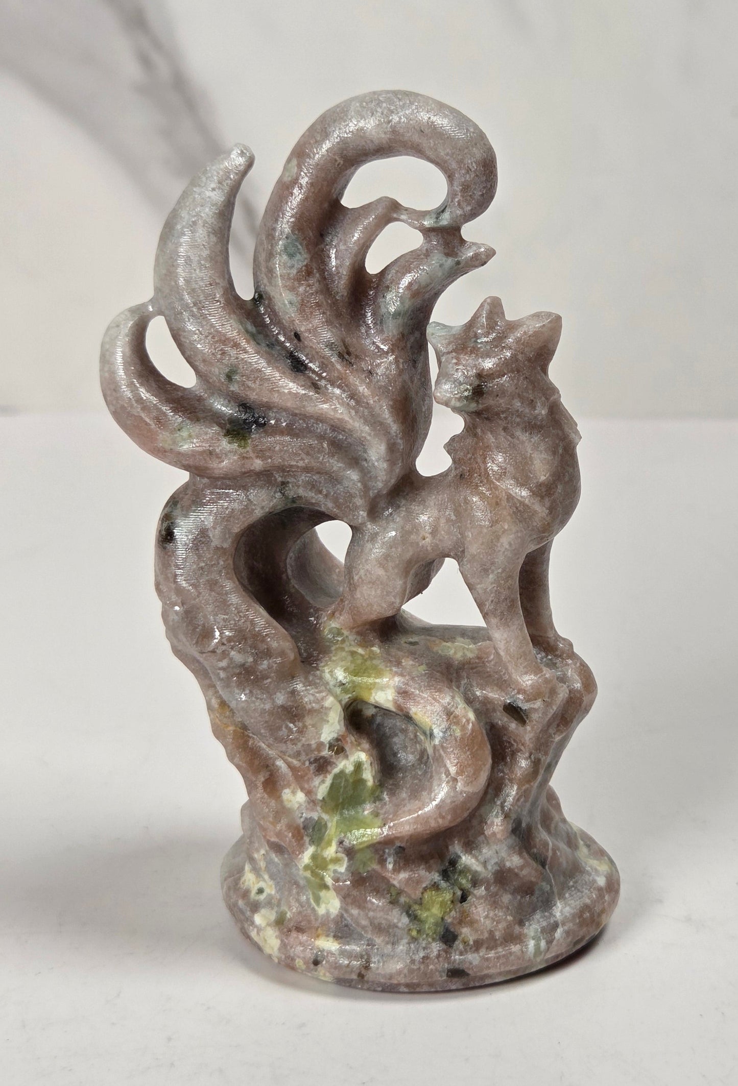 A small carved figurine of a fox with a flowing tail, standing on a textured base, photographed against a light marble-patterned background.