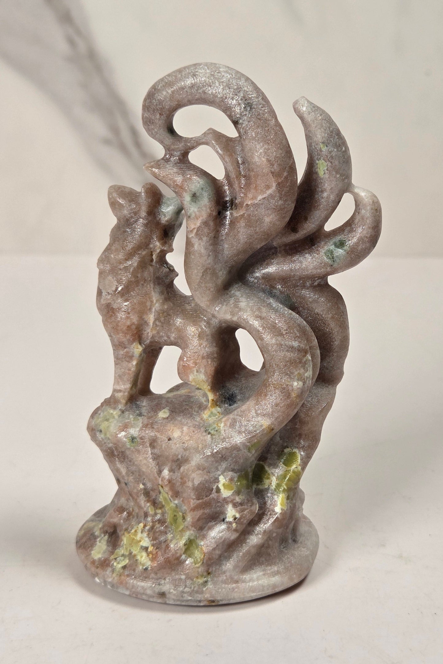 A small sculptural figurine of a fox-like creature with multiple curled tails, standing on a textured base. The object has a weathered, earthy finish with gray, brown, and muted green patina details, set against a light marble-patterned background.