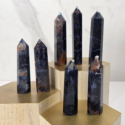 Seven polished crystal obelisks in dark blue, black, and brown marbled stone, displayed on a gold and light wood geometric pedestal arrangement.