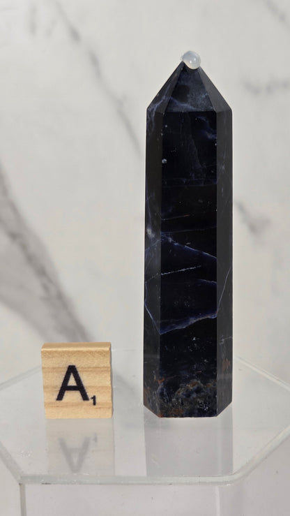 A tall, dark polished crystal point or tippice stone stands on a clear acrylic display base next to a small wooden letter tile showing the letter 'A'. The background is a soft-focus white and gray marble surface.