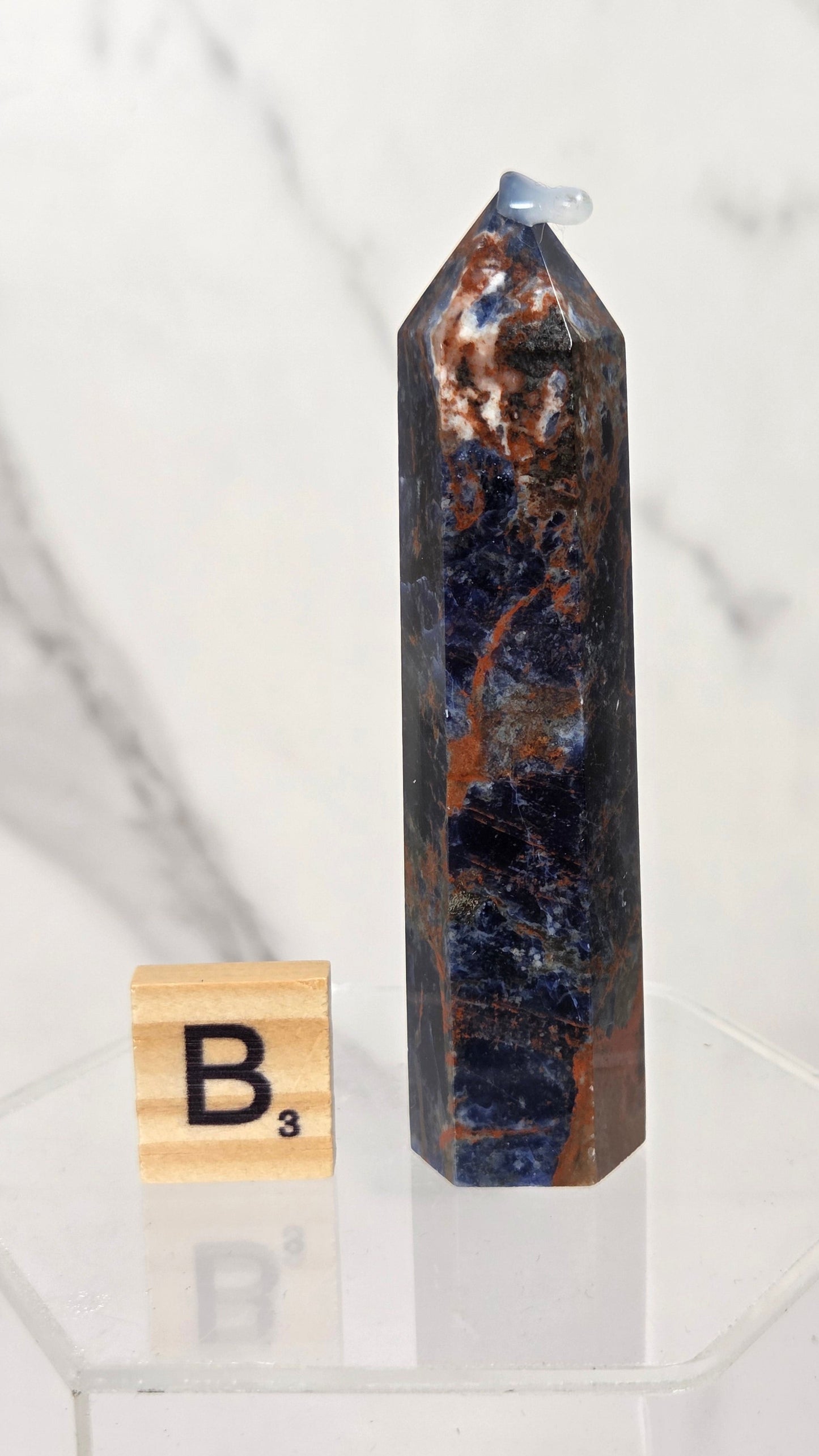 A tall, polished blue and copper jasper crystal point candle holder with a small white wick, displayed on a clear acrylic stand next to a wooden Scrabble tile showing the letter B.