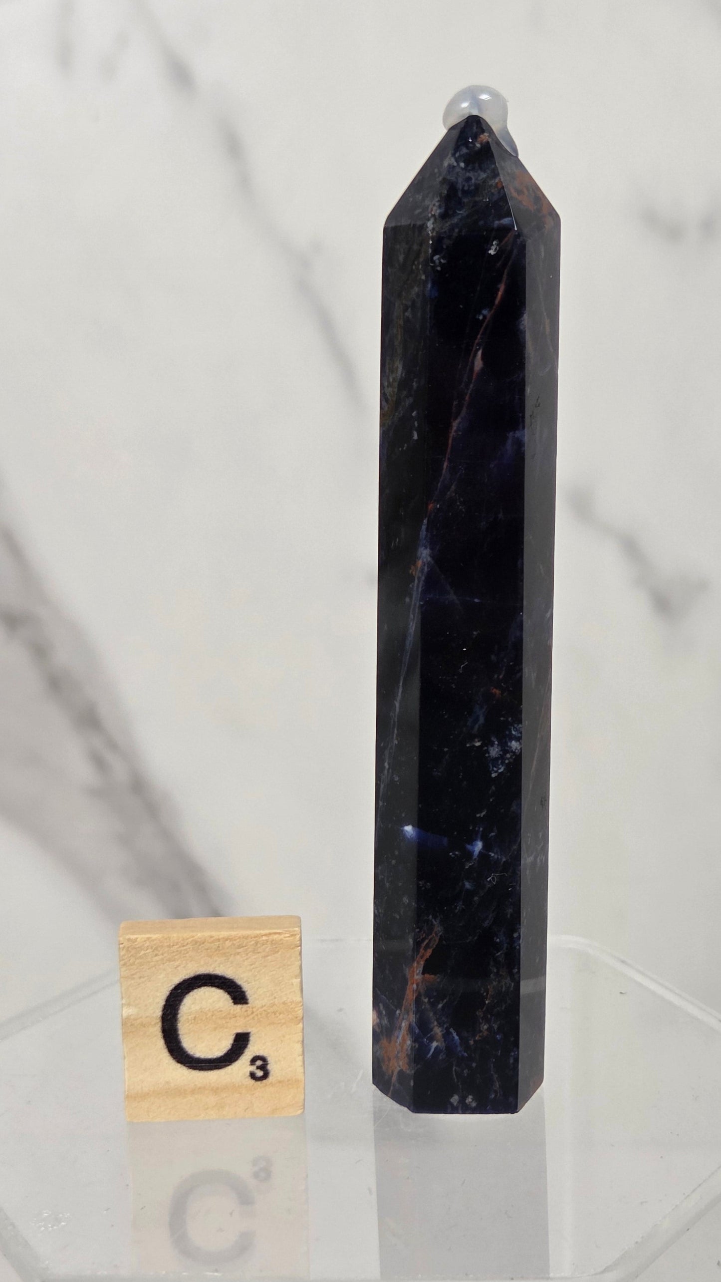 A tall, polished dark blue to black crystal point stands on a clear acrylic display base next to a small Scrabble tile marked C3, set against a light marble-patterned background.