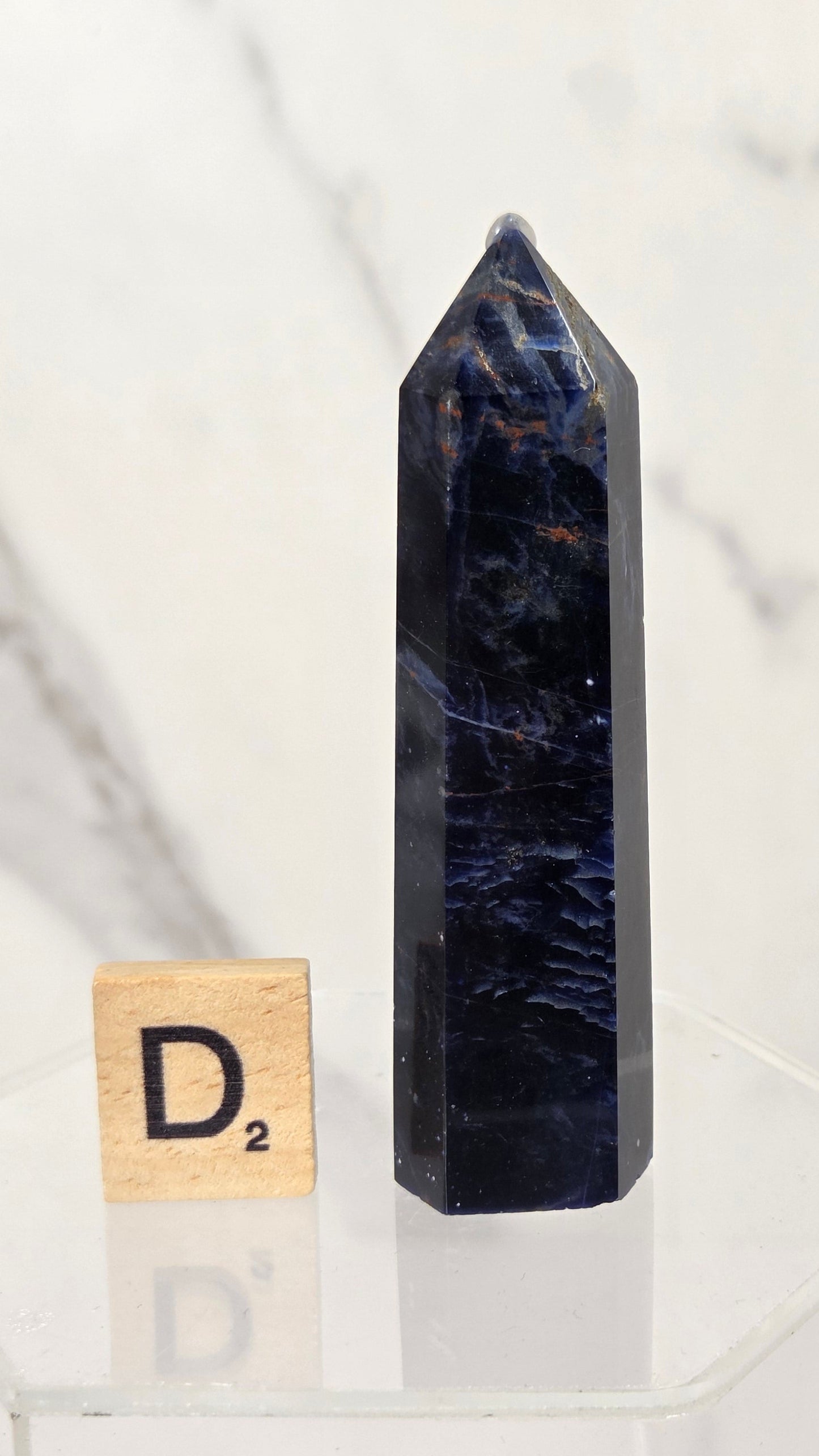 A tall, polished dark blue tuff crystal point tower stands on a clear acrylic display base next to a small wooden Scrabble tile showing the letter 'D'. The background is a soft-focus white and gray marble wall.