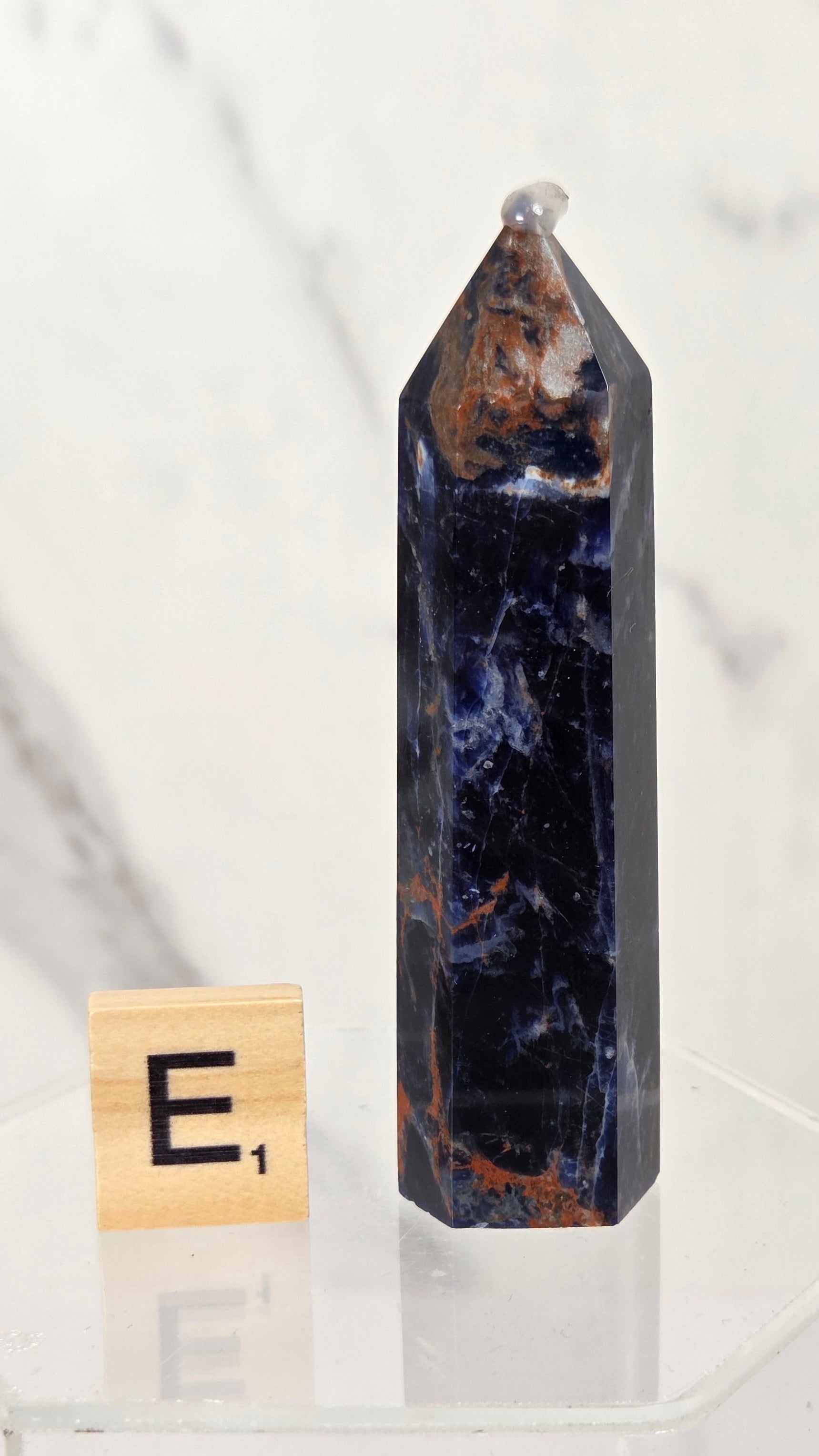 A polished dark blue and brown jasper crystal tower clogstone stands on a clear acrylic display base next to a wooden Scrabble tile showing the letter 'E'.