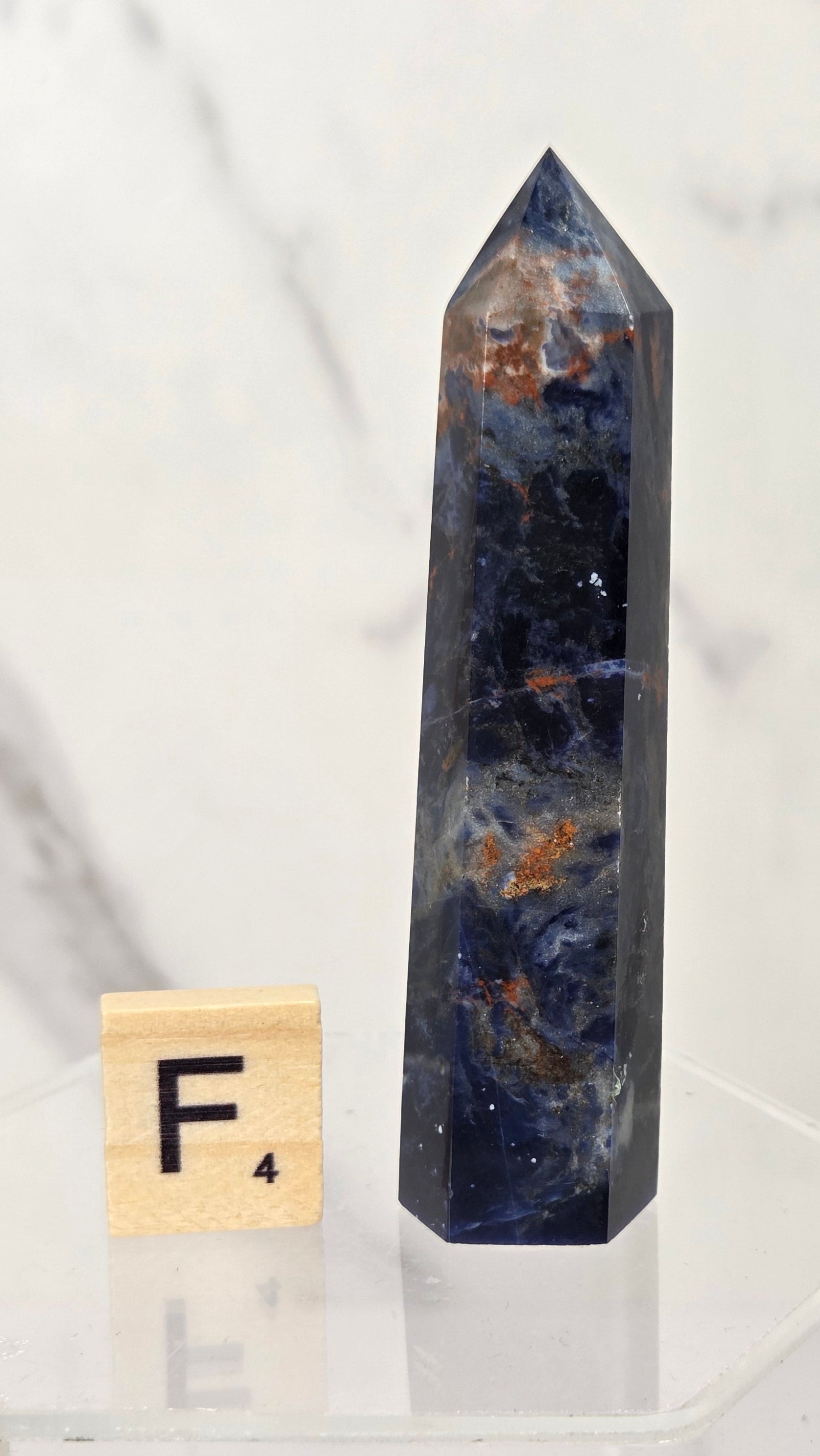 A polished blue selenite crystal tower with natural orange and white inclusions, displayed on a clear acrylic stand next to a wooden Scrabble tile showing the letter F.