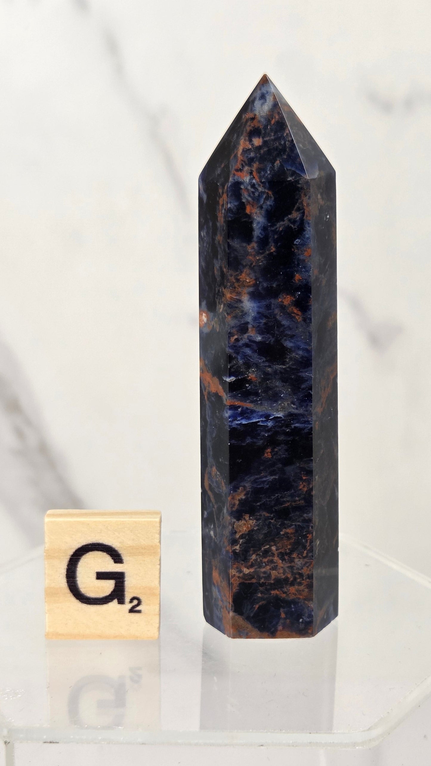 A tall, polished dark blue and black crystal point with brown veining stands on a clear acrylic display base next to a small wooden Scrabble tile showing the letter G.