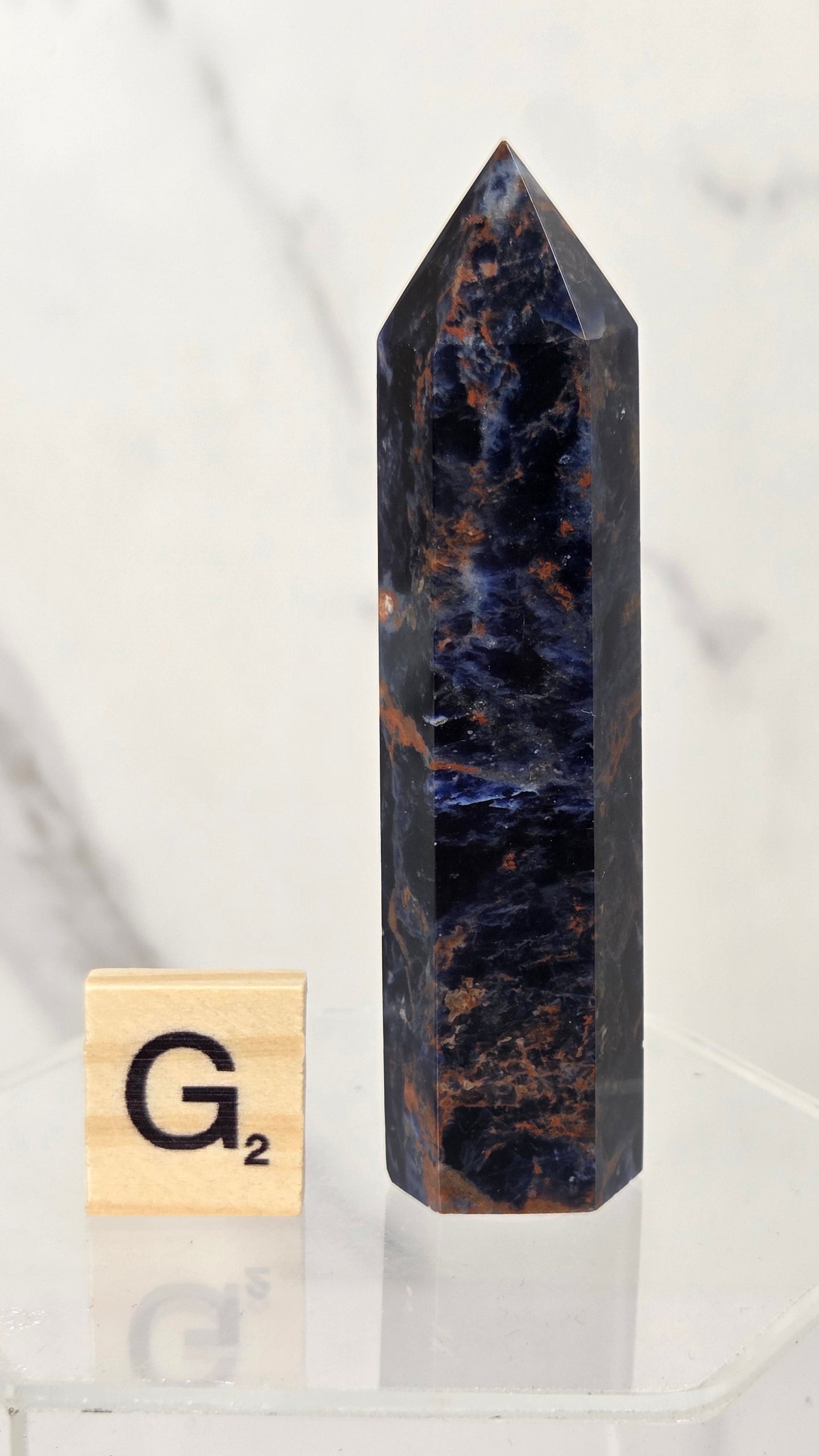 A tall, polished dark blue and black crystal point with brown veining stands on a clear acrylic display base next to a small wooden Scrabble tile showing the letter G.
