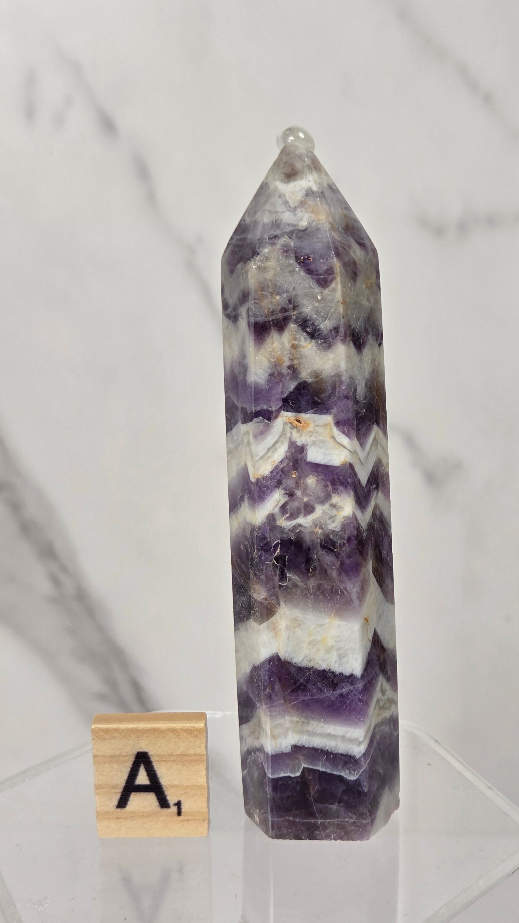 A pointed amethyst crystal tower on a clear acrylic display stand, next to a wooden Scrabble tile showing the letter 'A'. The crystal features natural purple, white, and gray banded patterns.