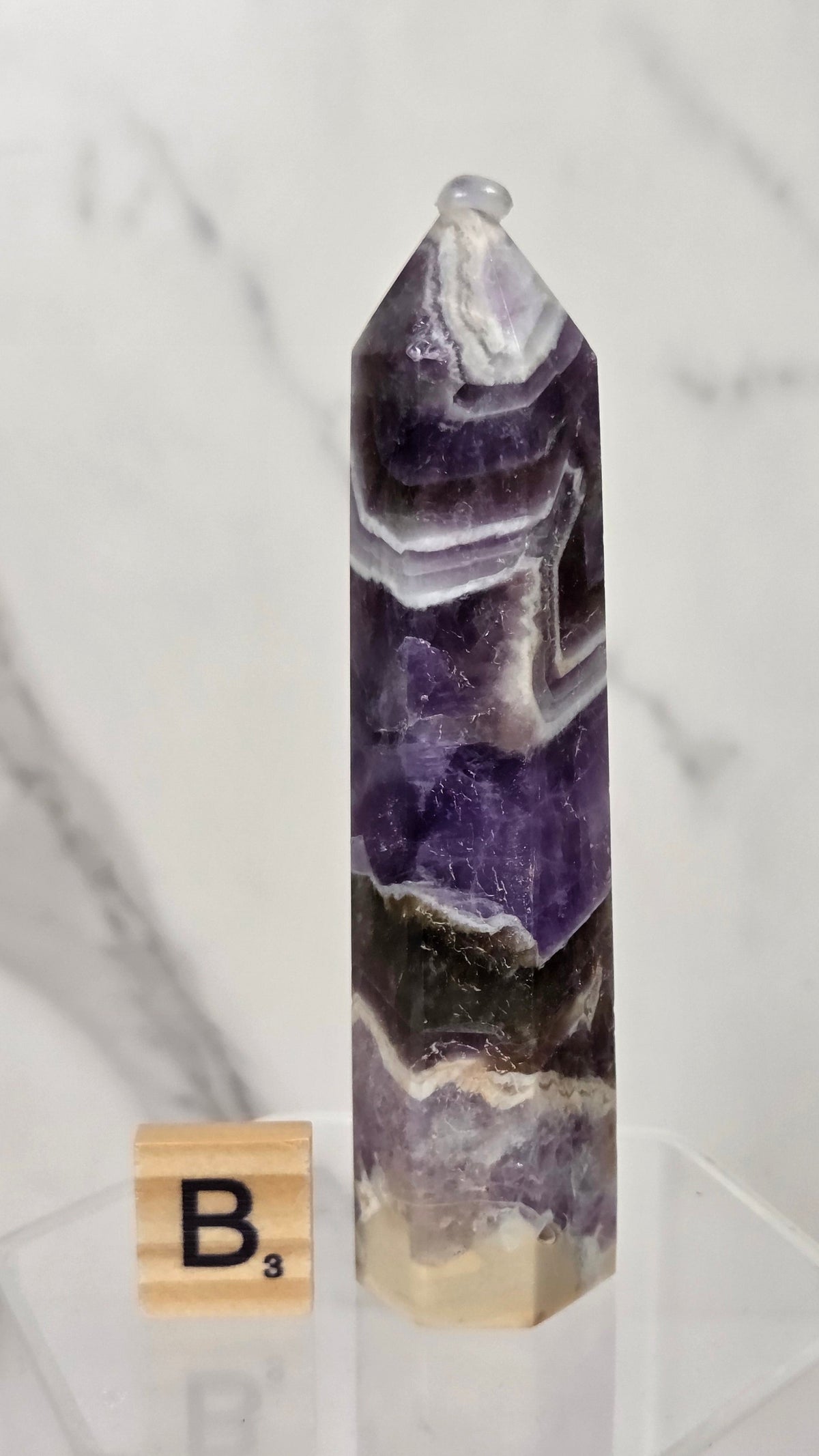 A polished amethyst crystal tower with natural banding in purple, white, and black, displayed on a clear acrylic stand next to a small Scrabble tile showing the letter B.