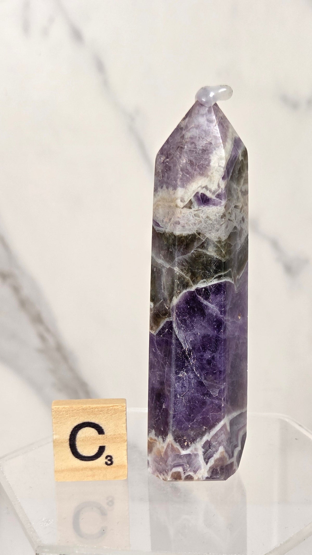 A natural amethyst crystal point with visible green and white matrix inclusions, displayed on a clear acrylic stand next to a Scrabble tile showing the letter C.