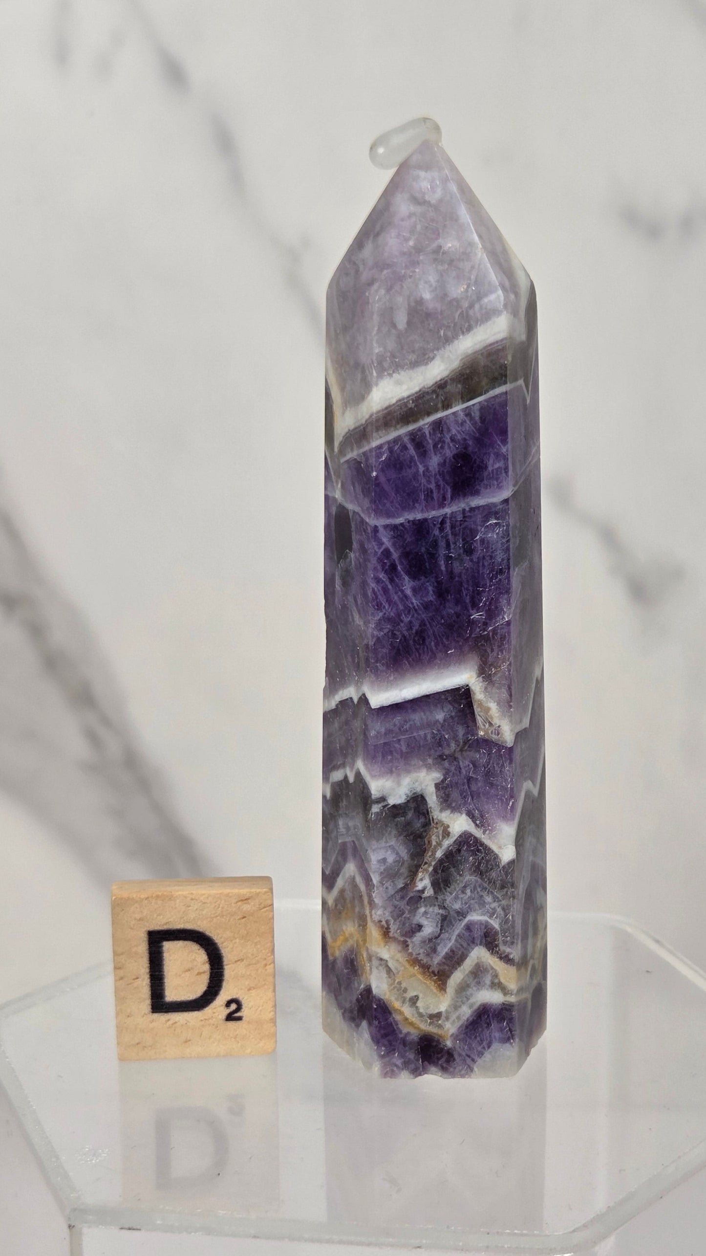 A pointed purple and white agate crystal tower on a clear acrylic display block, with a small wooden Scrabble tile showing the letter D² next to it, set against a white marble background.