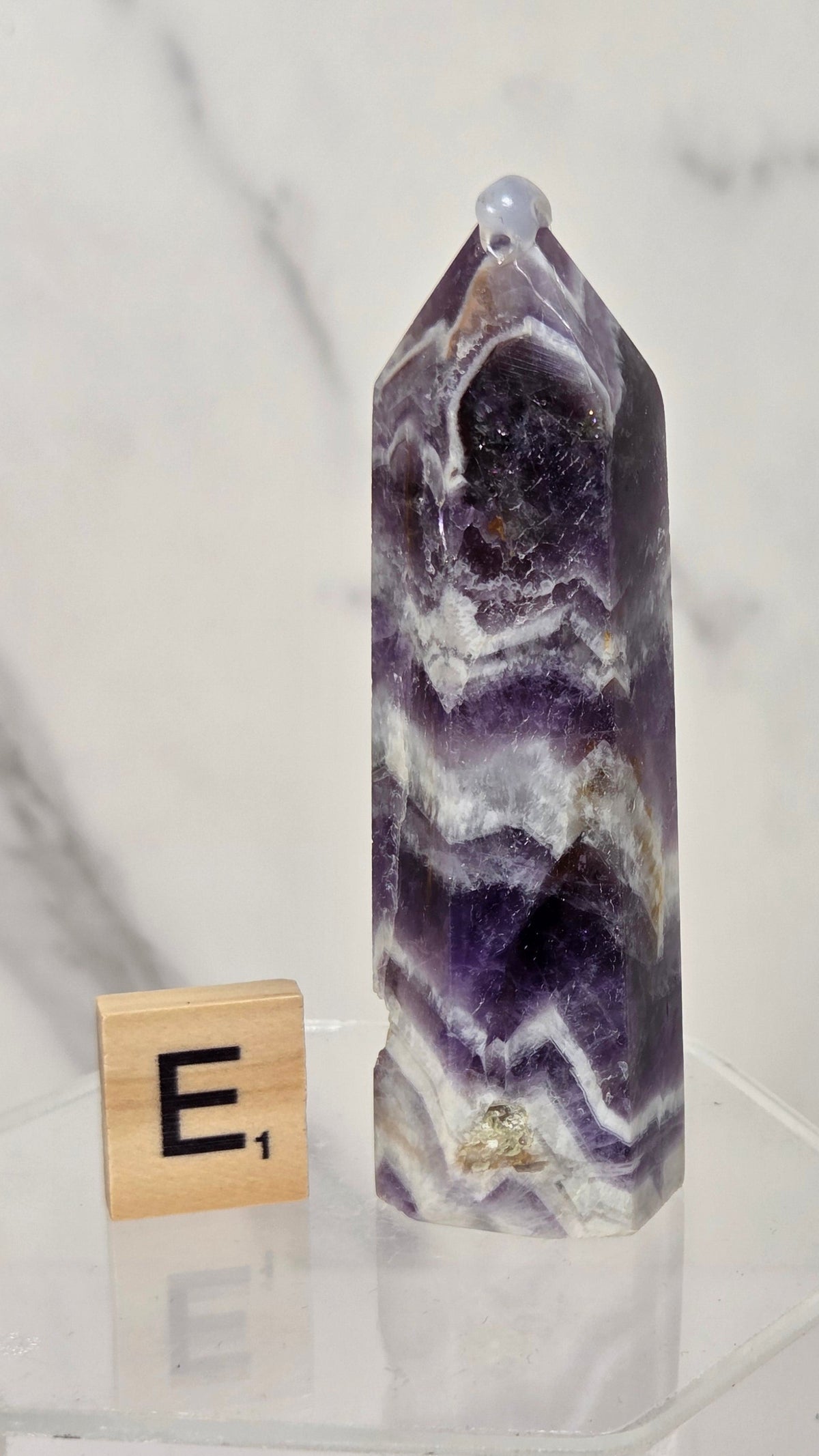 A polished purple amethyst crystal point tower with natural white and gray banded patterns, displayed on a clear acrylic hexagonal stand next to a wooden Scrabble tile showing the letter 'E'.