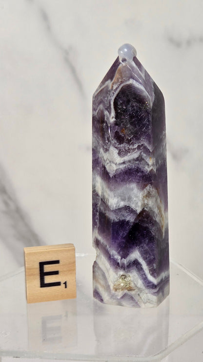 A polished purple amethyst crystal point tower with natural white and gray banded patterns, displayed on a clear acrylic hexagonal stand next to a wooden Scrabble tile showing the letter 'E'.
