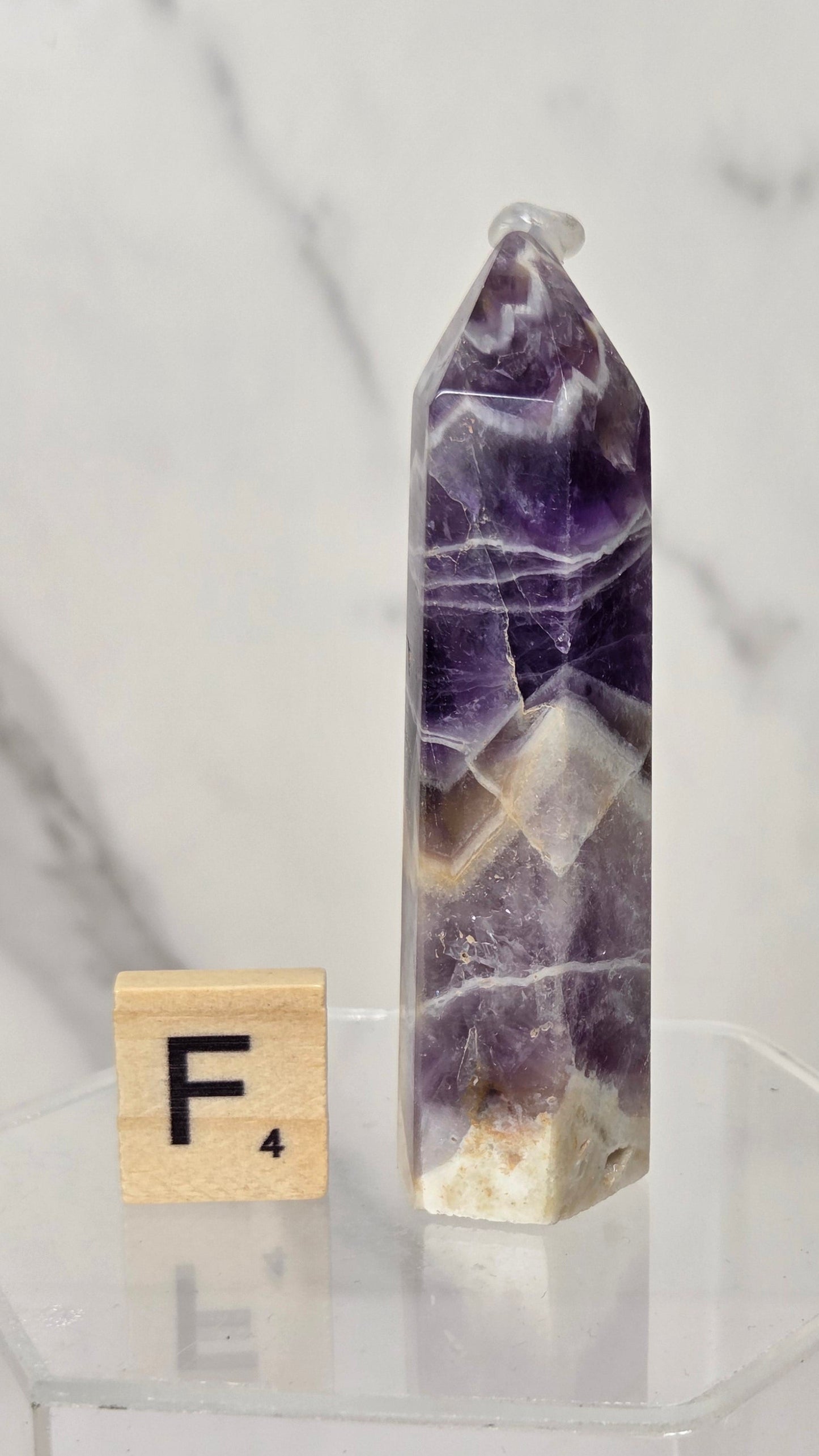 A polished purple amethyst crystal tower with natural white and beige banding, displayed on a clear acrylic pedestal next to a wooden Scrabble tile showing the letter F.