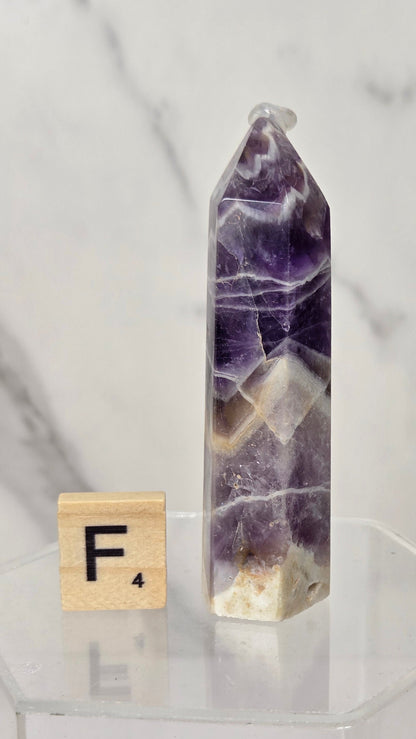 A polished purple amethyst crystal tower with natural white and beige banding, displayed on a clear acrylic pedestal next to a wooden Scrabble tile showing the letter F.
