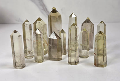 A group of natural pointed crystal points in translucent smoky gray and pale yellow tones, arranged on a light gray surface with a marble-patterned background.