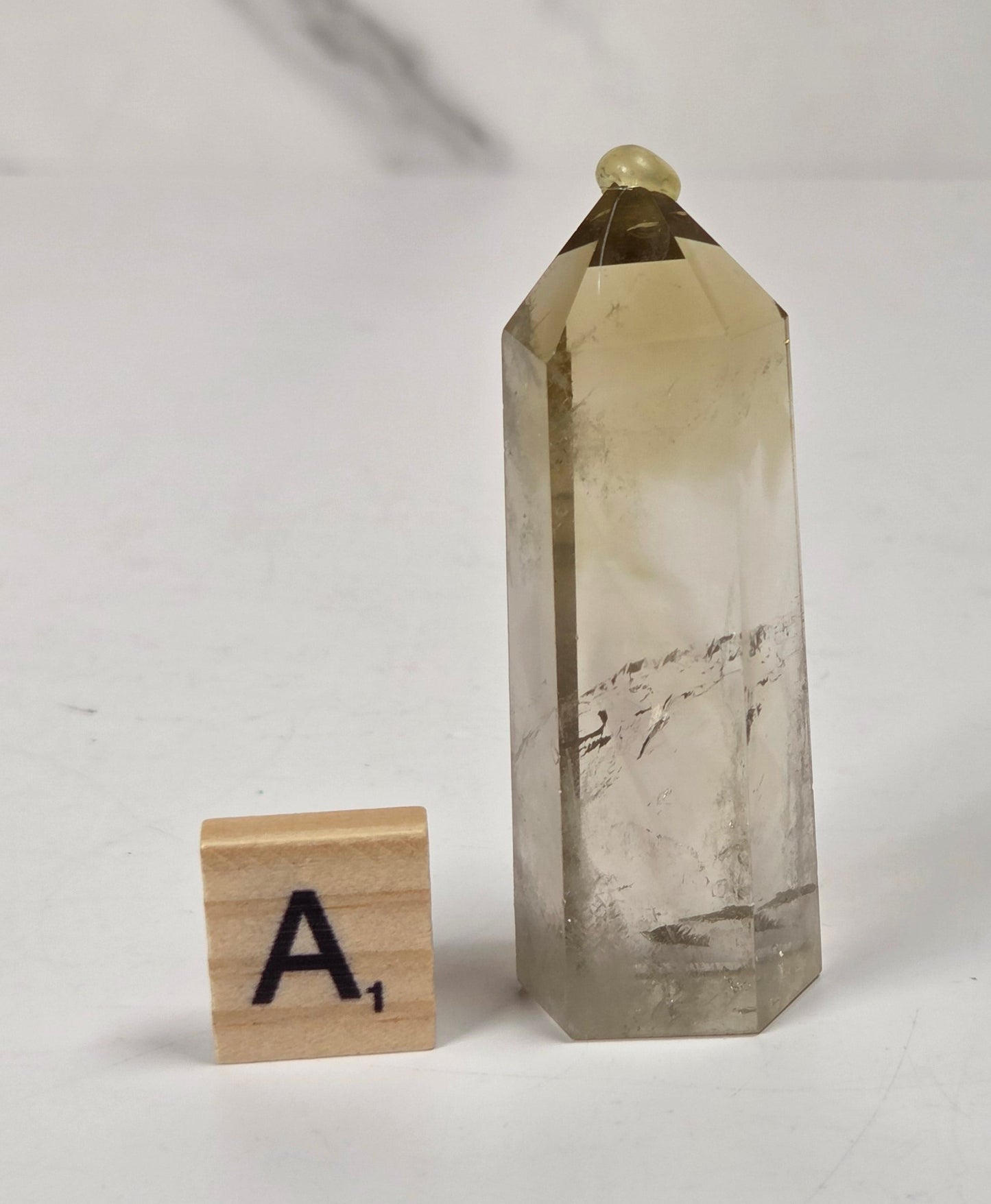 A natural smoky quartz crystal point with a small gold bead at its apex, displayed next to a wooden Scrabble tile on a white surface with a marble-patterned background.