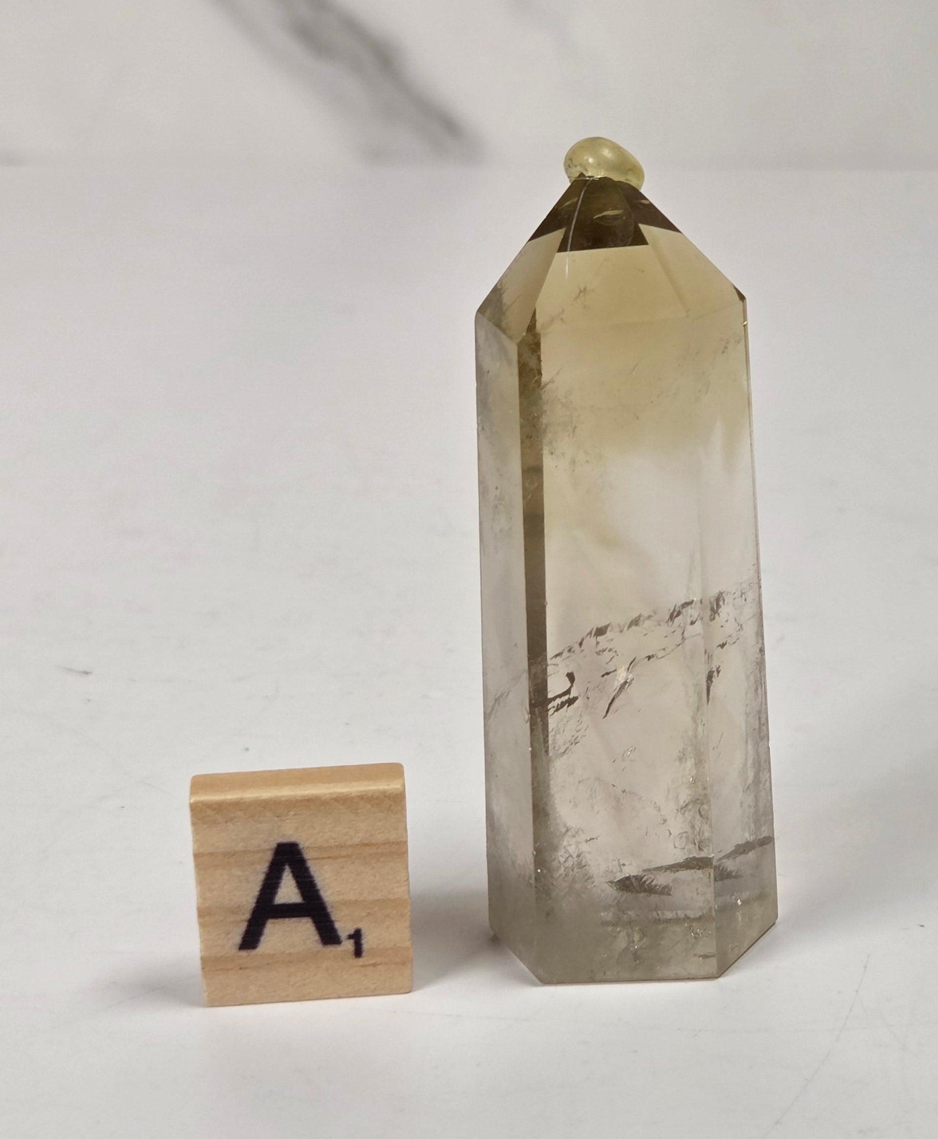 A natural smoky quartz crystal point with a small gold bead at its apex, displayed next to a wooden Scrabble tile on a white surface with a marble-patterned background.