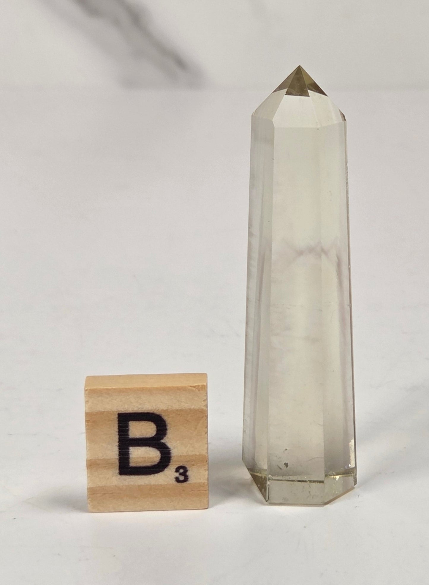 A tall, slender clear quartz crystal tower with a pointed tip stands on a white surface next to a small wooden Scrabble tile showing the letter B.
