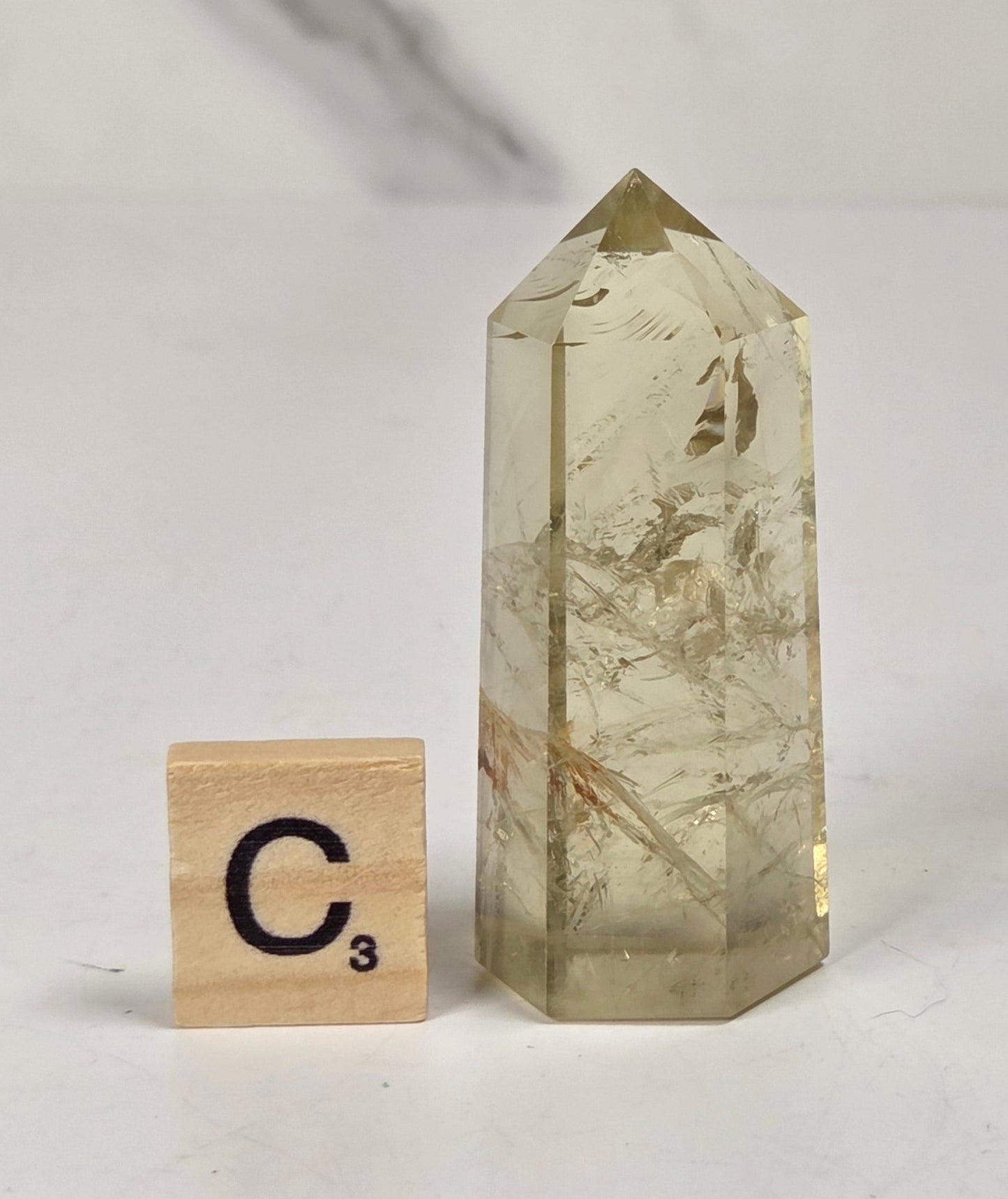 A tall, pointed clear crystal tower with internal beige and brown inclusions, standing on a white surface next to a small wooden Scrabble tile marked C3, against a light marble-patterned background.