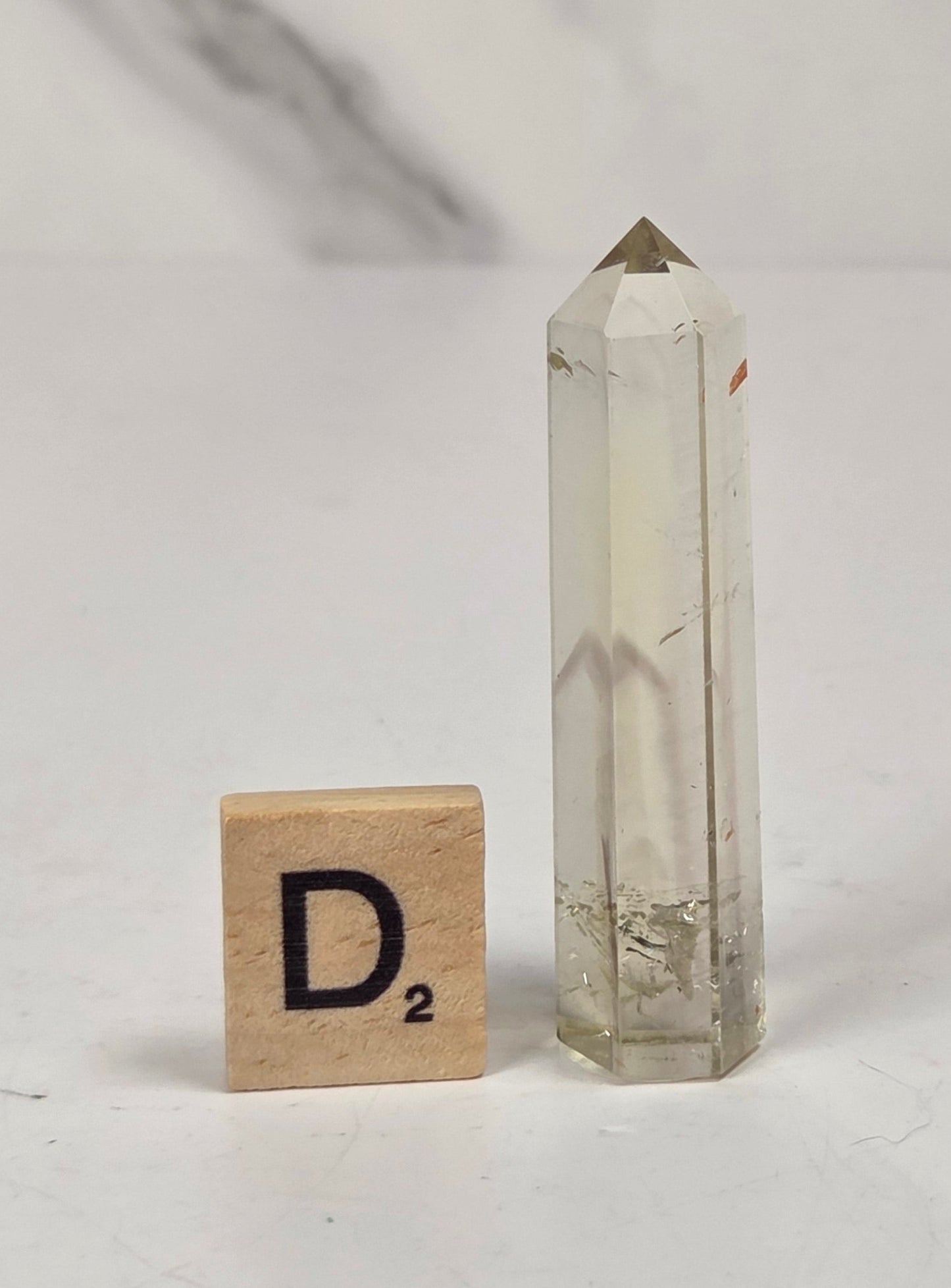 A clear quartz crystal point with a pointed metal cap stands next to a wooden Scrabble tile letter 'D' on a white marble-patterned surface.