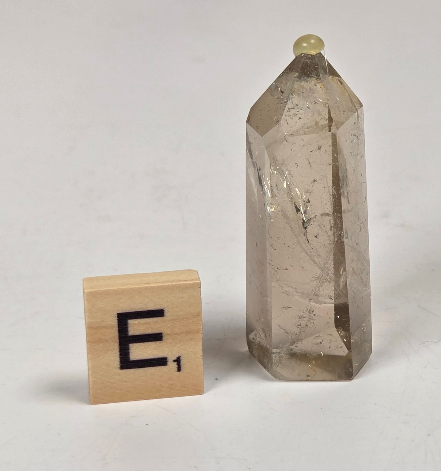 A small smoky quartz crystal tower with natural inclusions and a pointed top, shown next to a wooden Scrabble tile with the letter E on a light gray surface.