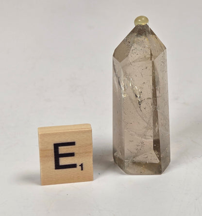 A small smoky quartz crystal tower with natural inclusions and a pointed top, shown next to a wooden Scrabble tile with the letter E on a light gray surface.