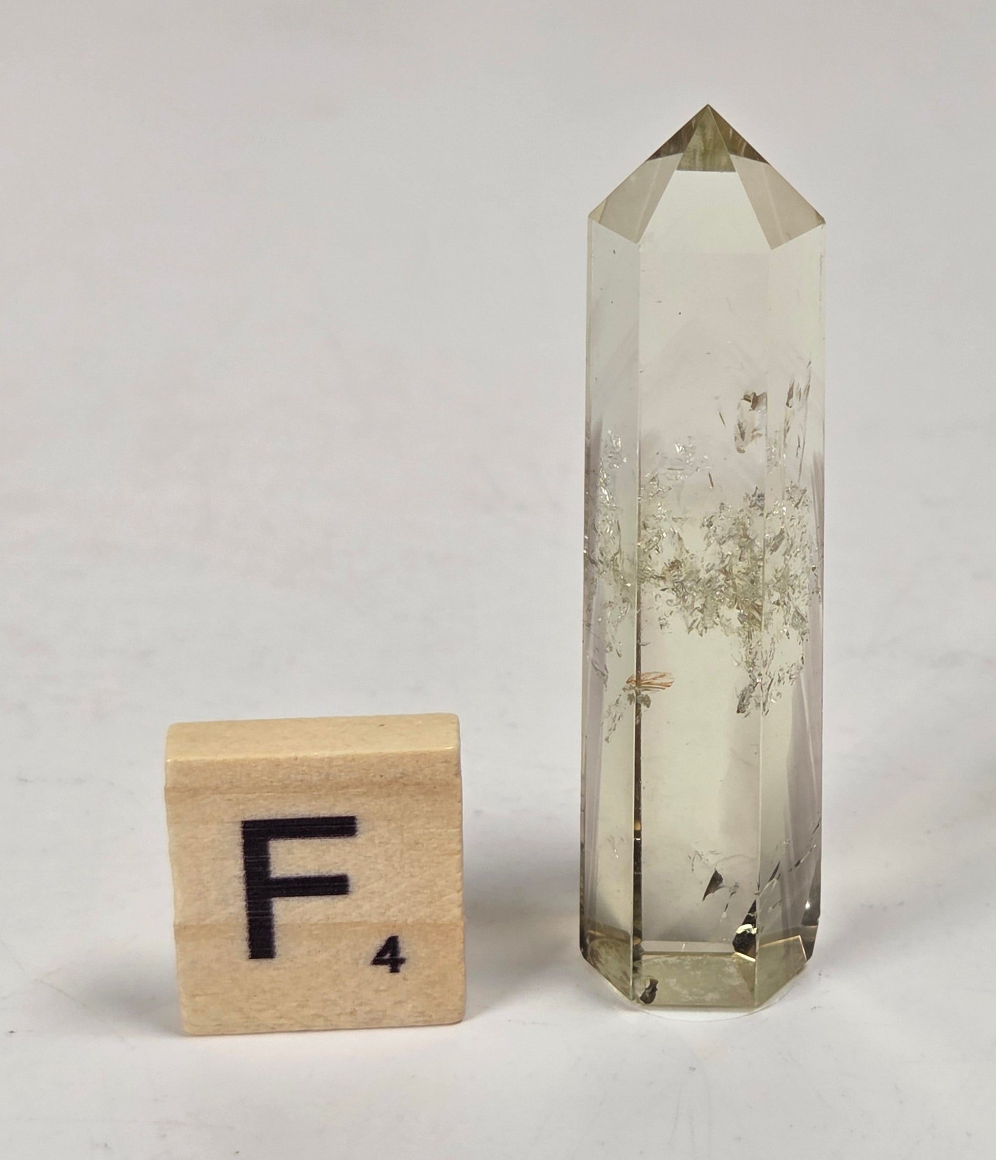 A tall, clear quartz crystal point with dried botanical inclusions, shown next to a wooden Scrabble tile spelling 'F4' on a light gray surface.