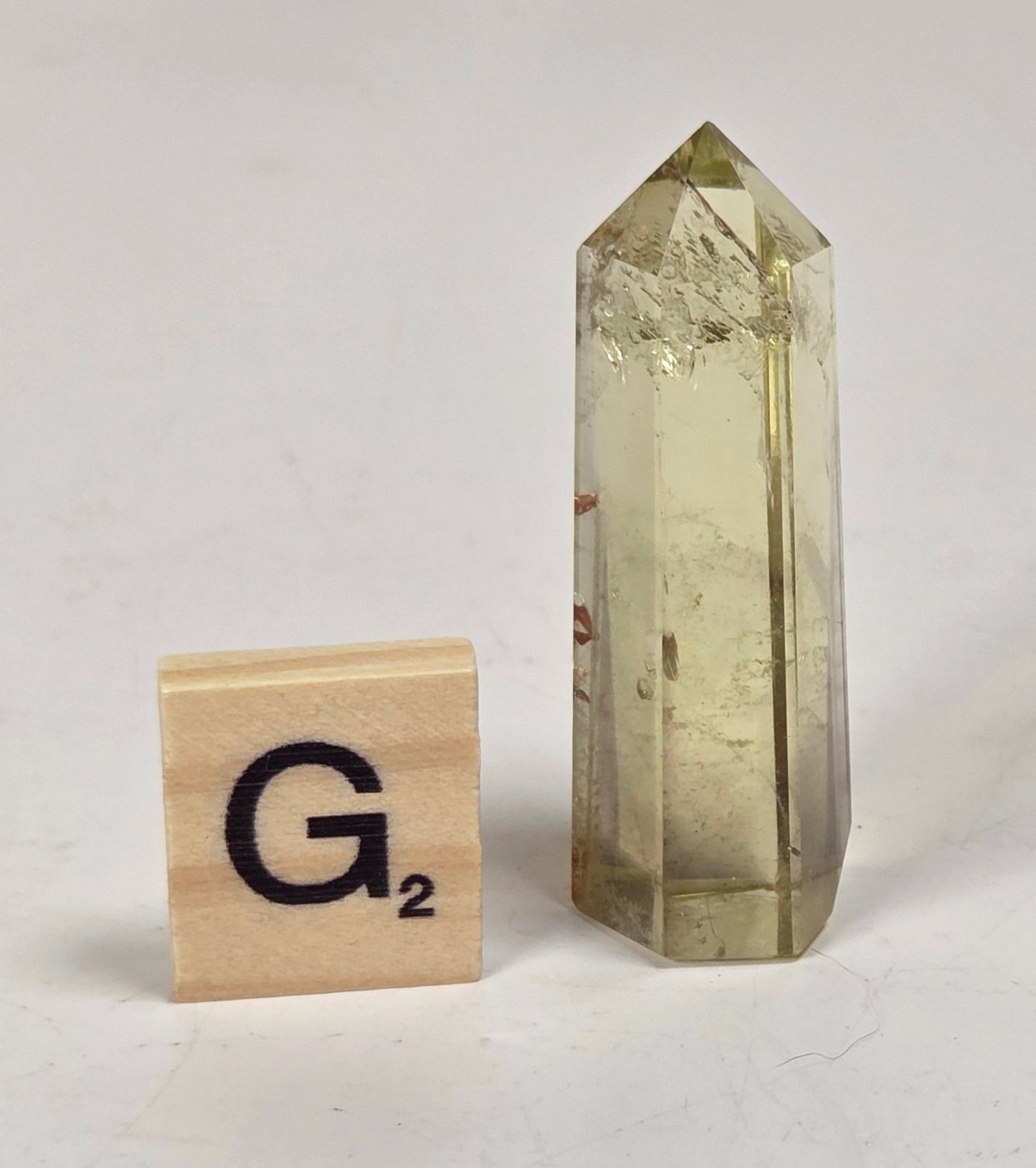 A tall, pointed clear crystal tower with a pale yellow-green tint stands on a white surface next to a wooden Scrabble tile showing the letter 'G'.