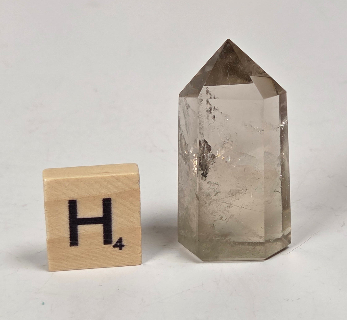 A natural smoky quartz crystal point with a pointed top and hexagonal base, shown next to a wooden Scrabble letter tile with the letter 'H' on a light gray surface.