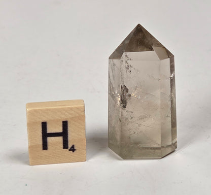 A natural smoky quartz crystal point with a pointed top and hexagonal base, shown next to a wooden Scrabble letter tile with the letter 'H' on a light gray surface.