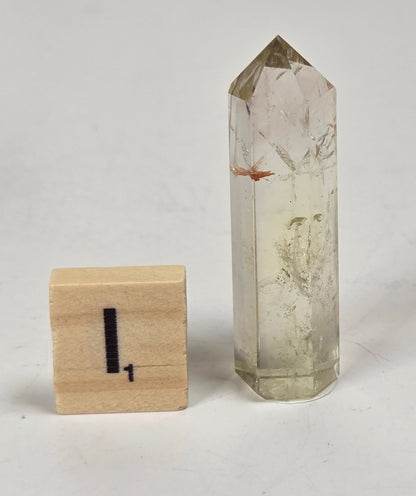 A natural clear quartz crystal point with internal inclusions stands next to a small wooden block marked with the number one, set against a plain light gray background.