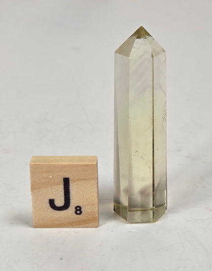 A clear pointed crystal tower with subtle yellowish-tinted internal bands, standing next to a small wooden Scrabble tile marked 'J' on a light marble-like surface.