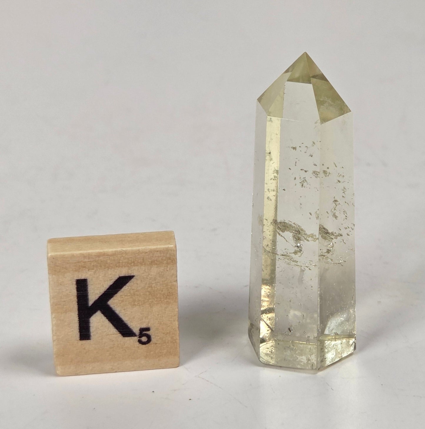 A clear yellow crystal point tower with natural inclusions, shown next to a wooden Scrabble tile spelling 'K' on a light gray marble-like surface.