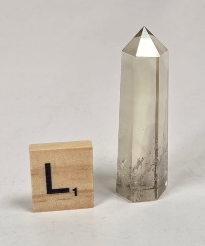 A natural clear quartz point crystal tower stands next to a wooden Scrabble tile showing the letter 'L' on a light gray marble surface.