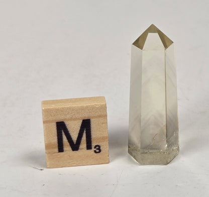 A clear faceted crystal point tower with a gold-toned pyramid cap, shown on a light marble-like surface next to a wooden Scrabble tile marked M³.